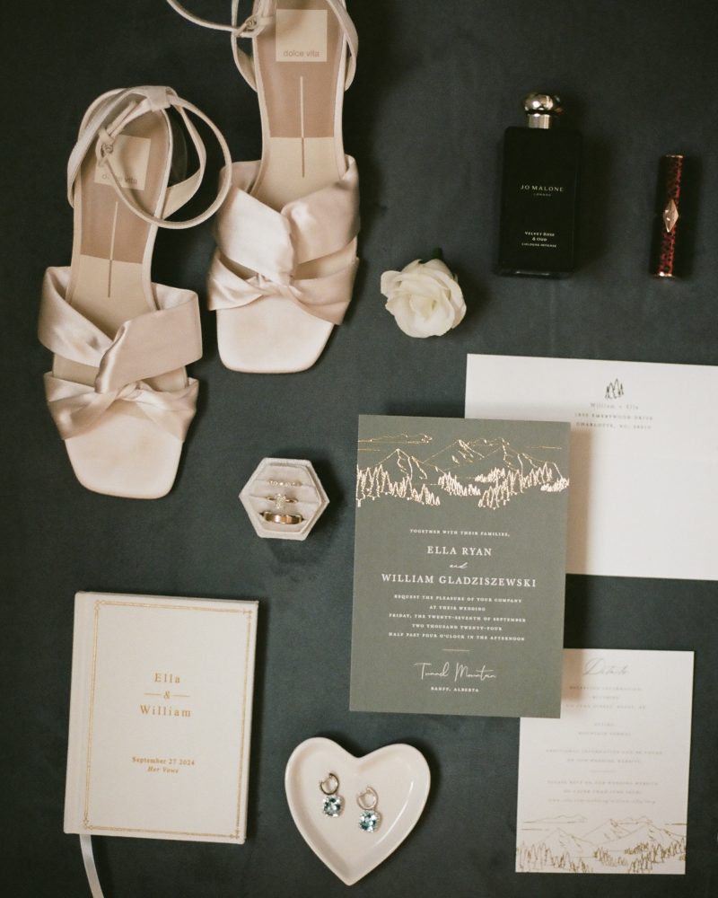 Flat lay of wedding items including white heeled sandals, a perfume bottle, lipstick, a white rose, rings in a box, earrings on a heart-shaped dish, invitation cards, and a white booklet—perfect inspiration for celebrations in Canmore or Banff.