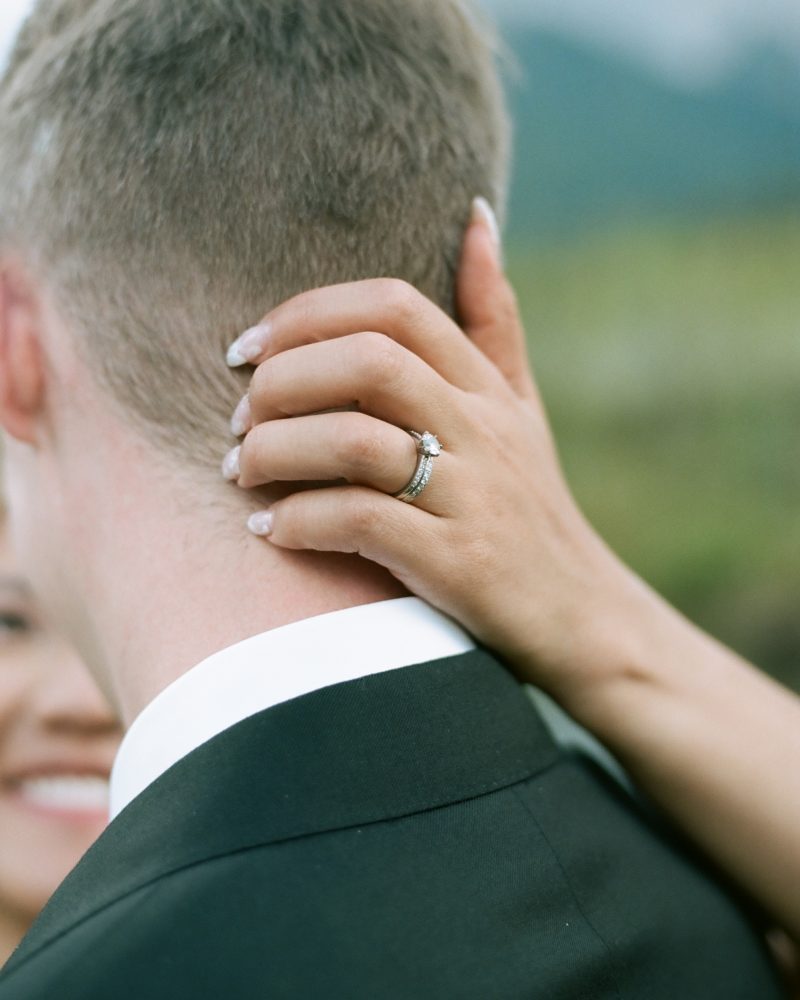 A close-up of a woman's hand with a diamond engagement ring resting on the back of a man's neck as they embrace outdoors in Banff, both dressed formally. The woman's face is partially visible, smiling—perfect for a wedding photographer's portfolio.