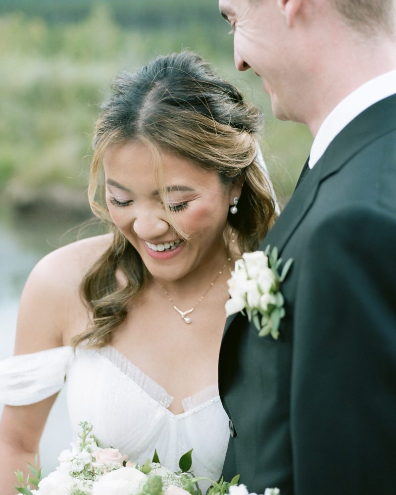 A bride in a white dress smiles and looks down, holding a bouquet of white and blush flowers, while standing close to her groom in a black suit with a boutonniere during their Banff wedding. They appear happy outdoors.