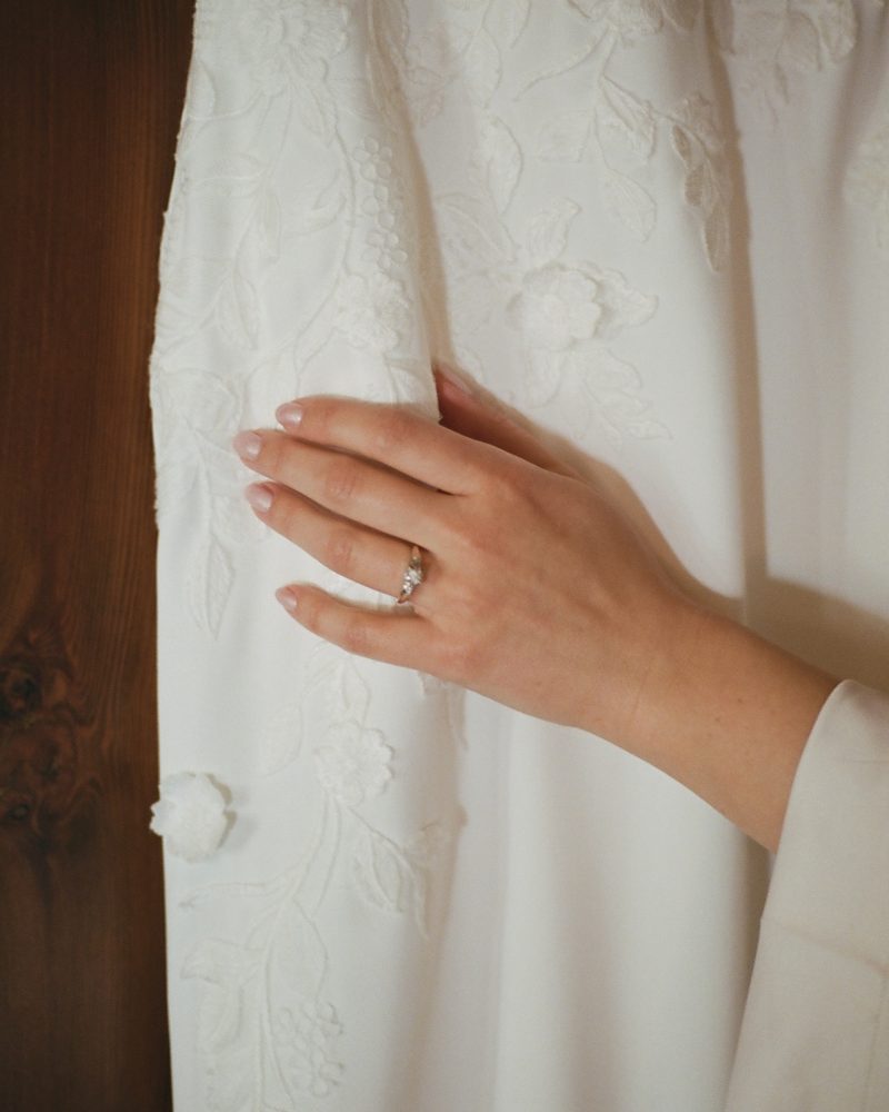 A hand with a French manicure and an engagement ring rests on a white, floral-embroidered wedding dress against a wooden background, capturing the elegance of a Calgary celebration.