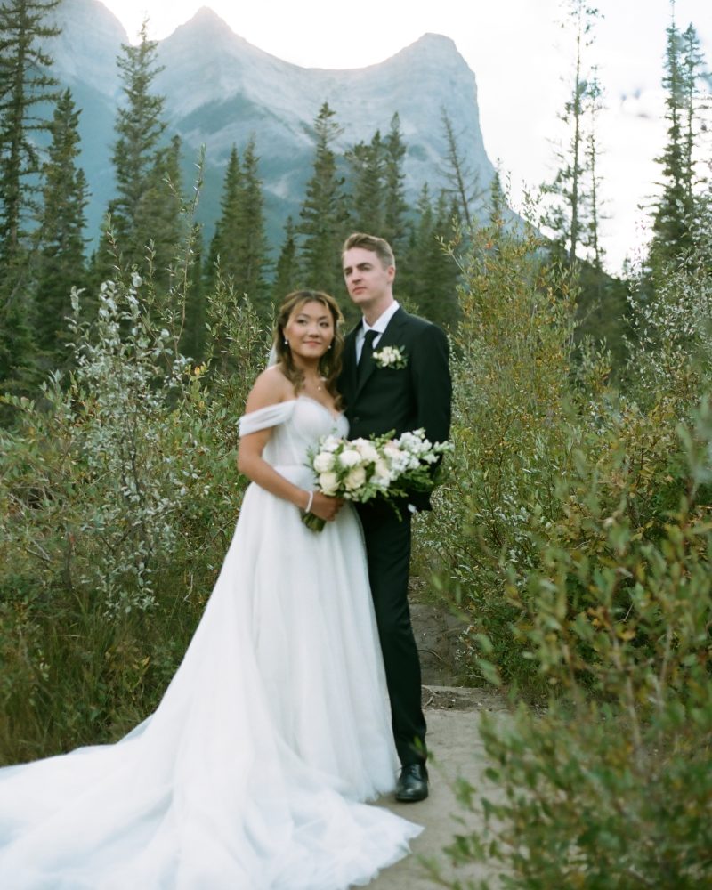 A bride in a white gown and a groom in a black suit stand together outdoors, surrounded by greenery and tall pine trees, with mountains visible in the background—a stunning Banff elopement. The bride holds a bouquet of flowers.