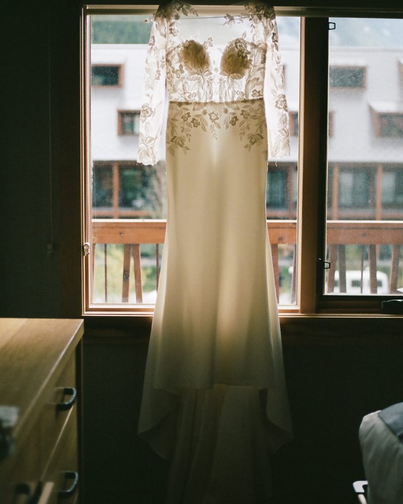 A long white wedding dress with lace detailing hangs in front of a window, sunlight shining through and Banff’s scenic buildings visible in the background.