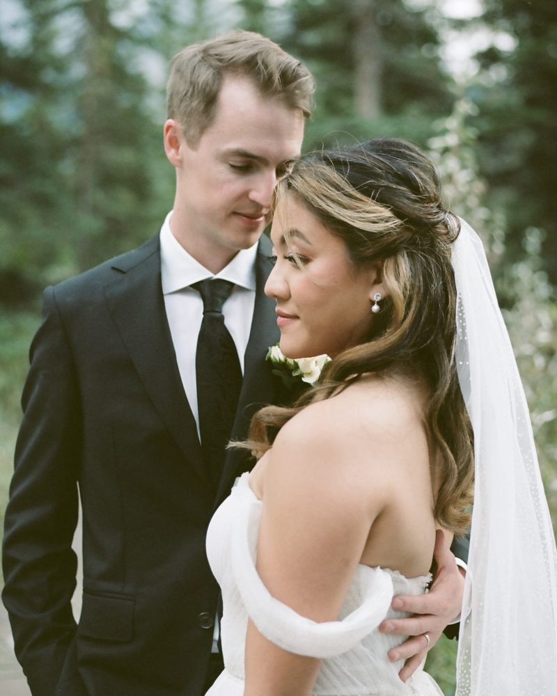 A bride and groom stand close together outdoors at their Calgary wedding. The bride, in an off-shoulder white dress and veil, faces sideways as the photographer captures the groom gently holding her waist amid pine trees.