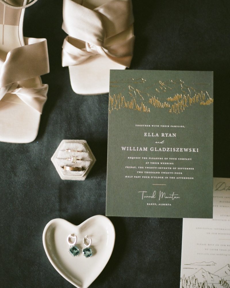 Flat lay by a wedding photographer: cream heeled sandals, a green and gold Canmore wedding invitation, a ring box with rings, emerald earrings on a heart-shaped dish, and another invitation on a dark fabric background.