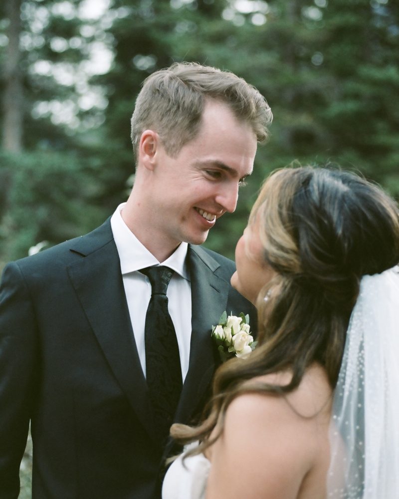 A bride and groom stand outdoors in Banff, facing each other and smiling warmly. The groom wears a black suit and tie, and the bride is in a white dress with a veil, set against a background of tall green trees.