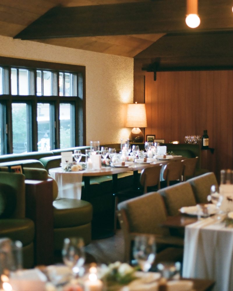 A warmly lit restaurant dining area in Canmore with tables set for a meal, featuring green chairs, white tablecloths, wine glasses, and modern hanging lights—perfect for an intimate elopement or cozy dinner near Calgary.