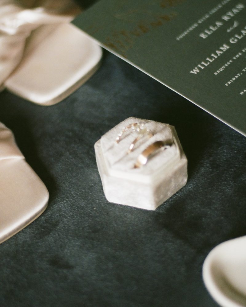 A pair of cream satin sandals, an invitation card, a hexagonal ring box with rings, and a heart-shaped dish with earrings are elegantly displayed on a dark surface—perfect details for a Calgary elopement or intimate wedding.