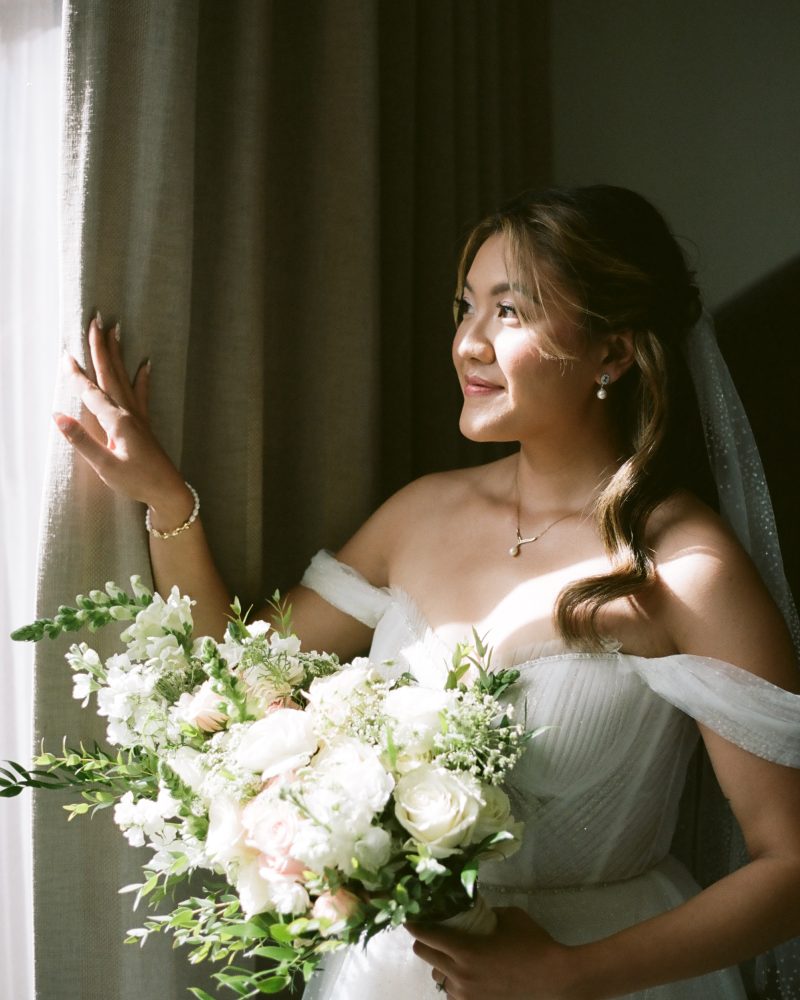 A bride in an off-shoulder white wedding dress holds a bouquet of white and pale pink flowers, standing by a window with beige curtains, softly lit by natural sunlight during her Canmore elopement.