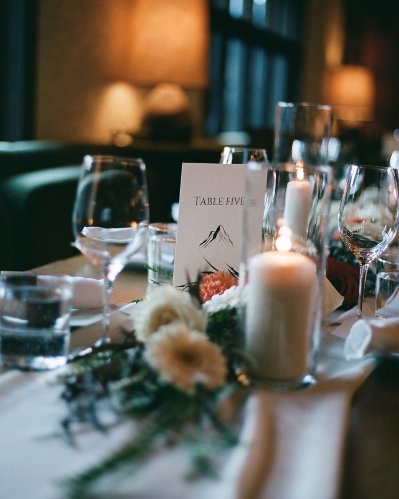 A softly lit table set for a Calgary wedding, with wine glasses, a burning candle, white flowers, greenery, and a card reading Table Five featuring a mountain illustration—perfect inspiration for any photographer.