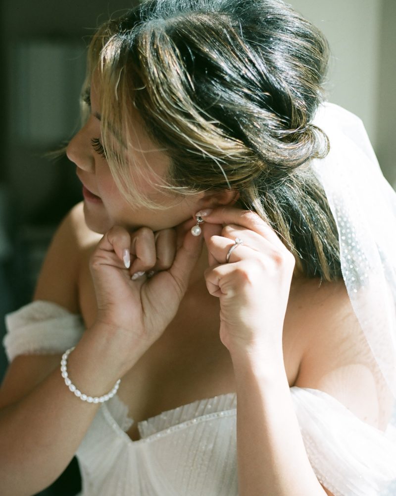 A bride in an off-the-shoulder white dress adjusts a pearl earring during her Banff elopement, with a matching bracelet on her wrist and a veil in her hair.