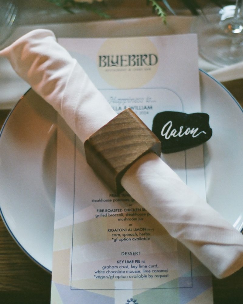 A rolled white napkin with a wooden ring and a name card labeled Devon rests on a plate atop a printed Bluebird menu, set on a wooden table in Canmore, with a drink, lit candle, and photographer snapping moments nearby.