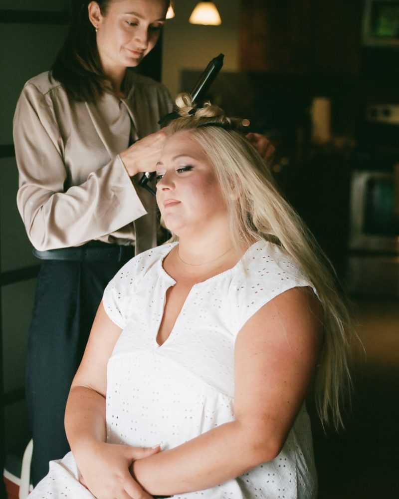 A woman with long blonde hair, wearing a white dress, sits with her eyes closed while another woman, possibly a wedding photographer in Canmore, styles her hair with a curling iron in a softly lit room.