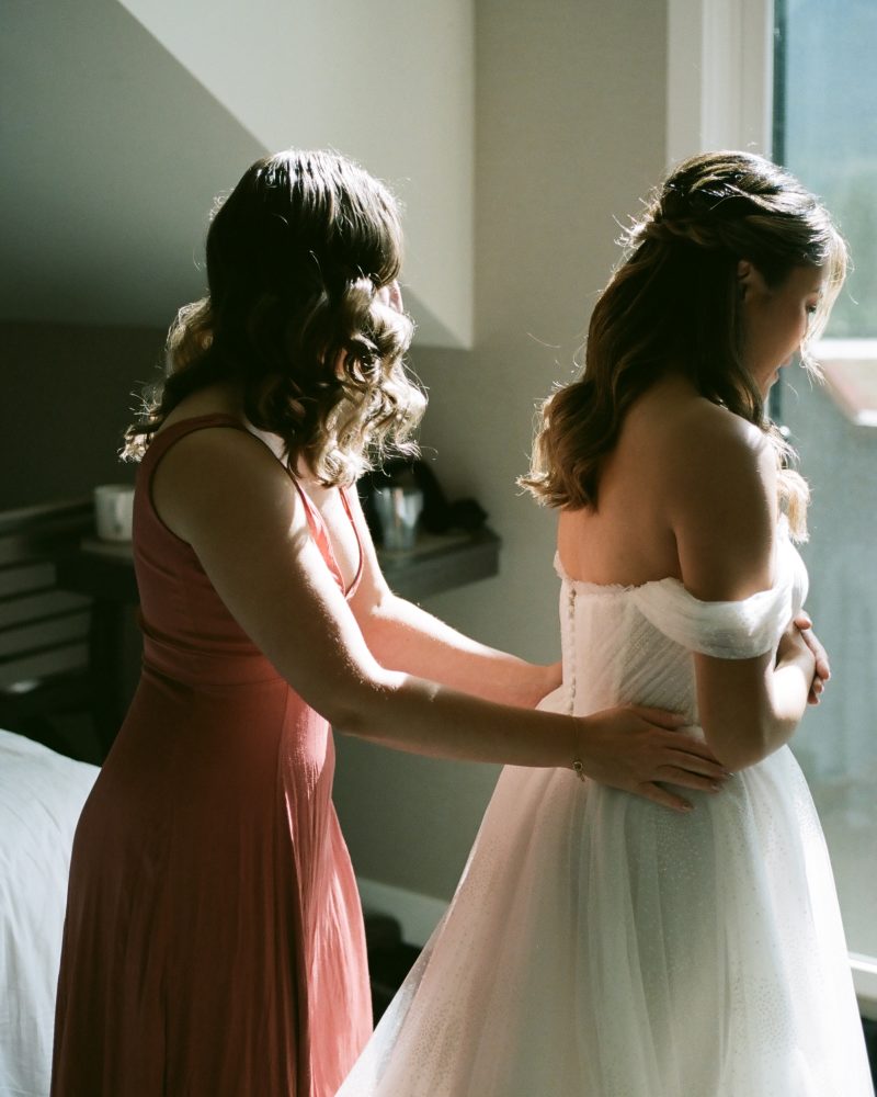 A photographer in Canmore captures a woman in a pink dress helping a bride adjust her off-shoulder wedding gown near a sunlit window, with soft natural light illuminating this intimate elopement moment.
