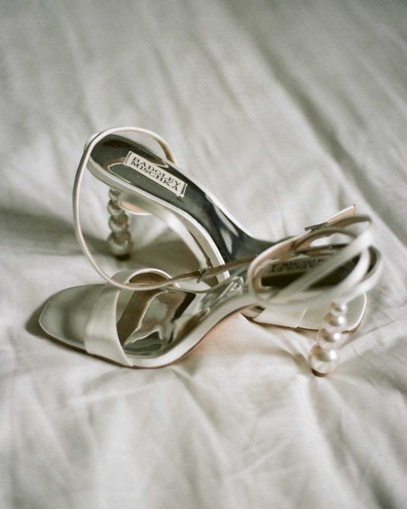 A pair of elegant white high-heeled sandals with pearl-adorned heels rests on a soft, white fabric background, perfect for a Calgary wedding or any photographer seeking timeless bridal detail shots.