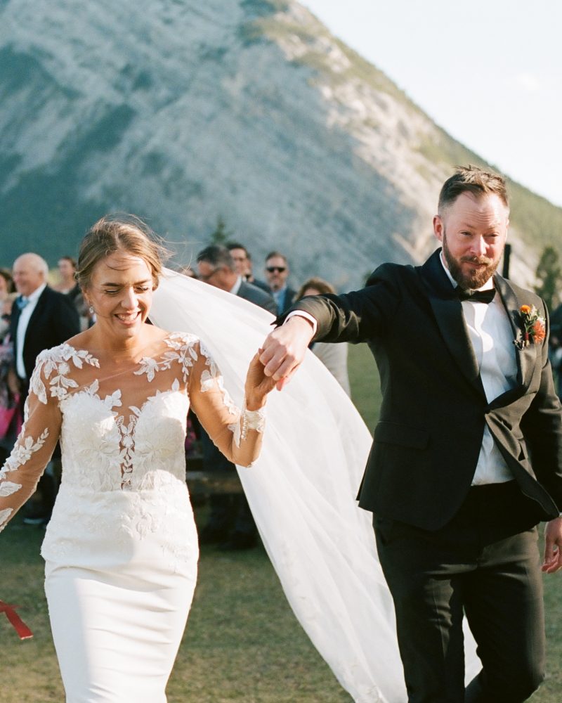 A bride and groom walk hand in hand outdoors after their Canmore wedding ceremony, smiling and joyful, with mountains and guests in the background. The bride wears a lace dress and veil, holding a bouquet.