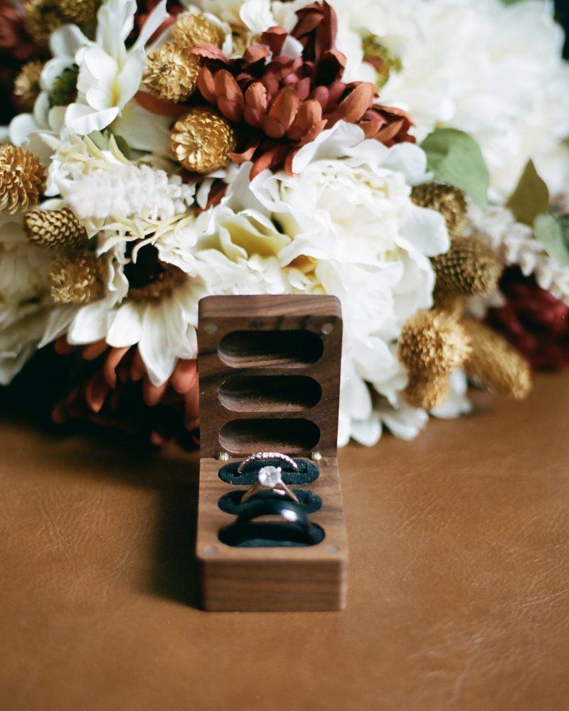 A wooden ring box displays three rings, including a diamond engagement ring, in front of a bouquet of white and orange flowers—perfect for a Calgary elopement—on a brown surface.