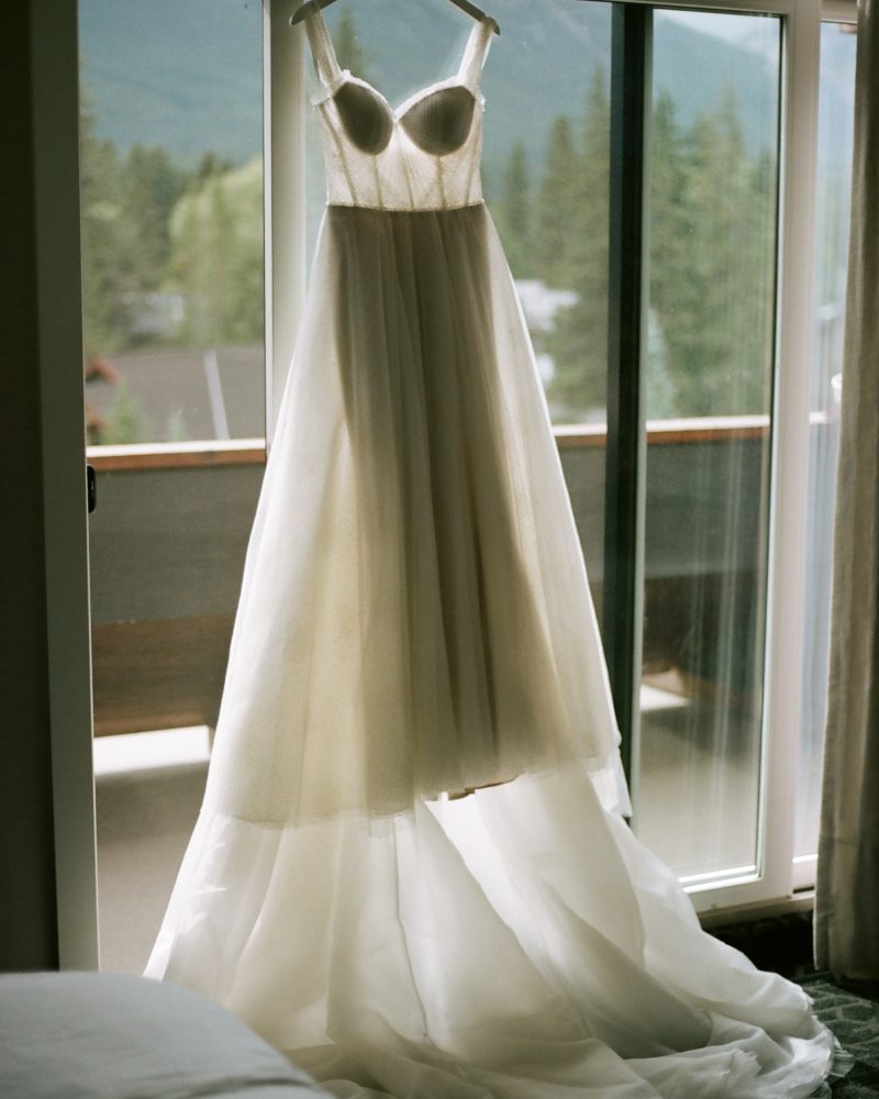A white, strapless wedding dress with a full tulle skirt hangs on a hanger in front of a glass sliding door, with trees and Banff mountains visible outside in the background—perfect inspiration for any Calgary photographer.