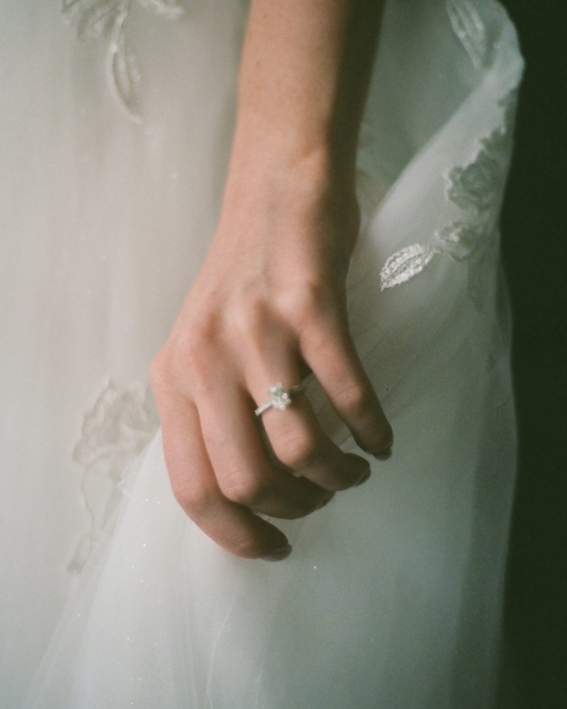 A close-up of a hand wearing a diamond ring rests against white, floral-embroidered fabric, likely part of a wedding dress. Soft lighting highlights the delicate details—captured beautifully by a Calgary elopement photographer.