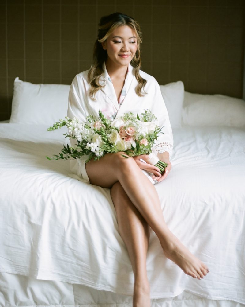 A woman sits on a white bed in a robe, smiling gently and holding a bouquet of white and pink flowers on her lap. Captured by a Calgary photographer, the scene features a neutral-colored wall and soft pillows in the background.
