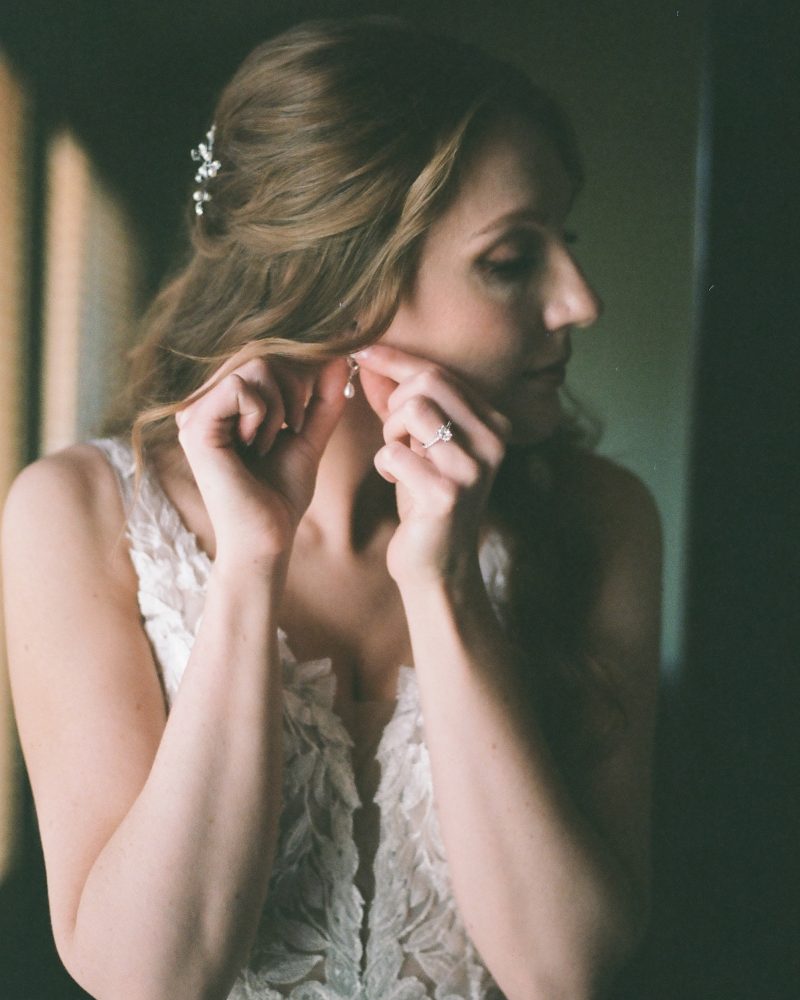 A bride with wavy hair in a white lace dress adjusts her earring before her Canmore elopement. She is facing sideways in soft lighting, with a delicate hair accessory and an engagement ring visible on her hand.