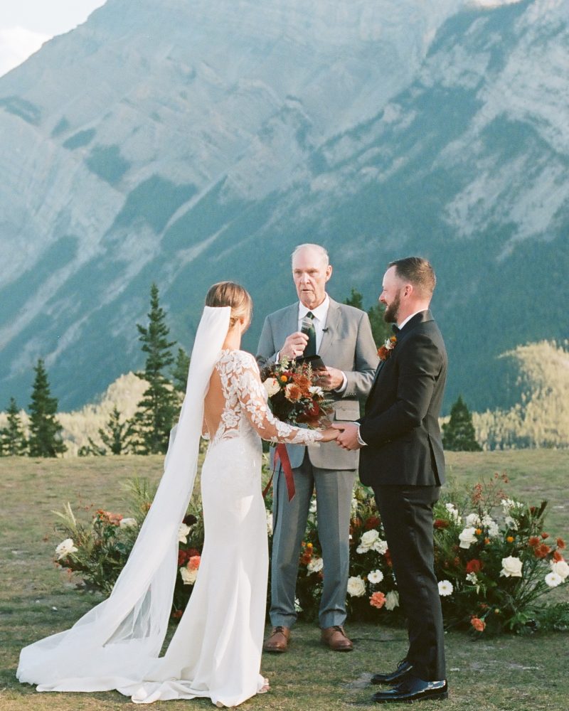 A bride and groom stand facing each other during an intimate Calgary elopement ceremony, with an officiant and attendants beside them. Mountains and trees create a scenic backdrop captured by the photographer under a partly cloudy sky.