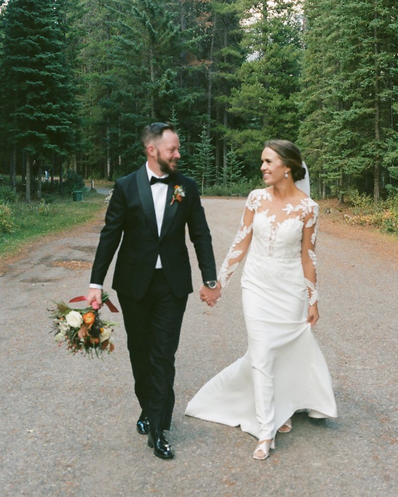 A bride and groom walk hand in hand down a forested path during their Canmore elopement. The groom wears a black tuxedo and holds a bouquet; the bride’s white lace-sleeved dress glows as they smile at each other, surrounded by trees.