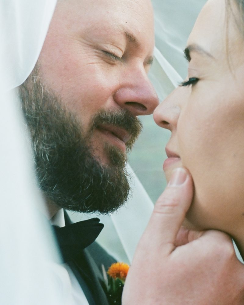 A close-up of a bride and groom under a veil, eyes closed, gently touching faces in an intimate moment. Captured by a Banff photographer, the bearded groom smiles softly while the serene bride in earrings radiates calm.