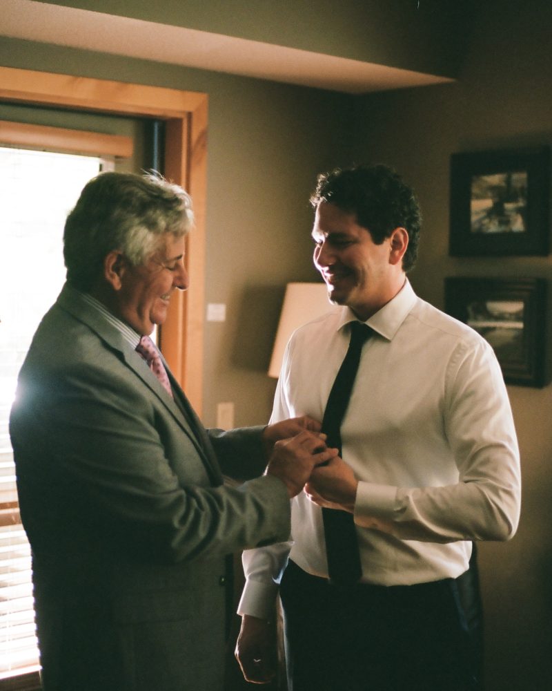 Two men in formal attire smile at each other indoors during a Calgary wedding. One, older with gray hair in a suit, helps adjust the younger man’s cufflinks. Warm light from a nearby window bathes the cozy scene.