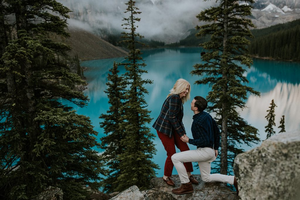 A man proposes on one knee to a woman on a rocky ledge near Calgary, surrounded by pine trees and overlooking a turquoise lake with mountains in the background—a perfect moment for an adventurous elopement.