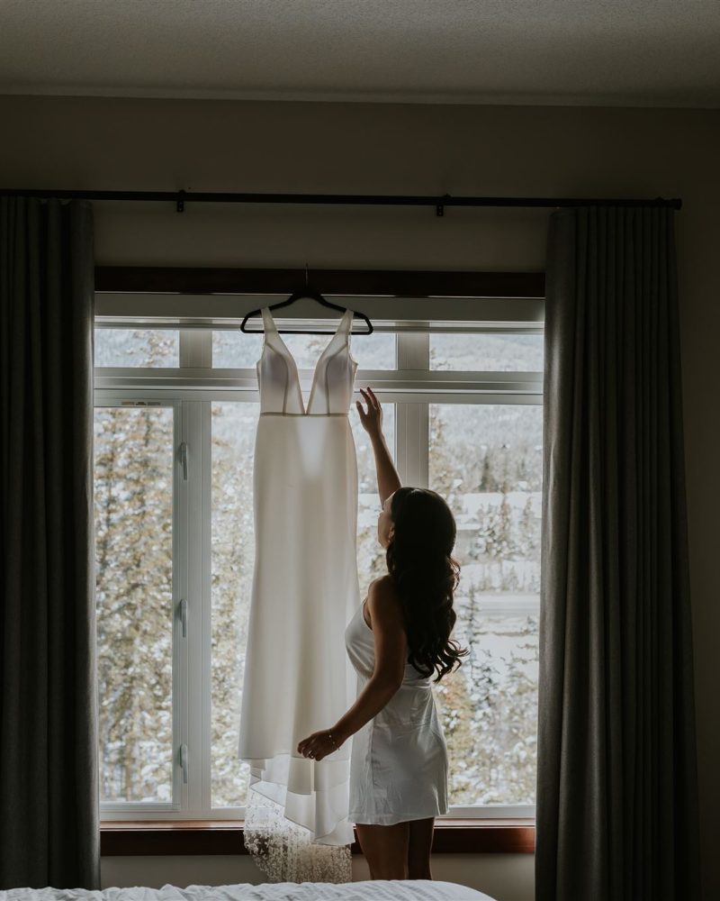A woman in white pajamas stands in a dimly lit bedroom, reaching up toward a white dress hanging in front of a large window with closed curtains. Snow-covered trees reminiscent of Banff are visible outside, capturing the eye of any photographer.
