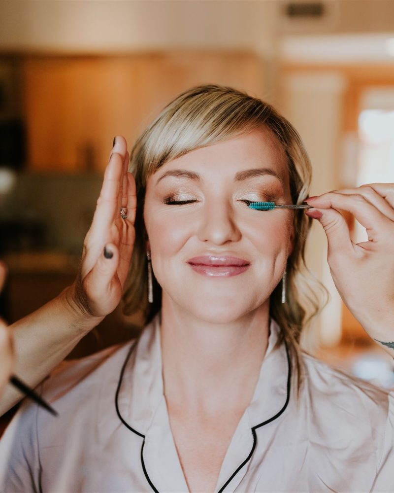 A woman with closed eyes smiles as a makeup artist applies bright blue eyeshadow to her eyelid. A photographer captures the moment in warm indoor lighting, likely preparing her for a special event or wedding in Banff.