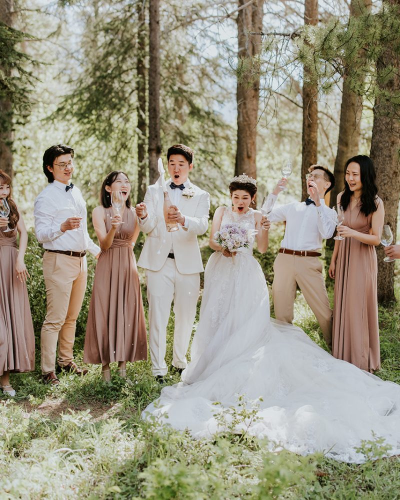 A bride and groom stand in the center of a forest near Canmore with four bridesmaids in mauve dresses and three groomsmen in white shirts and tan pants, all smiling for their photographer and holding drinks under the trees.
