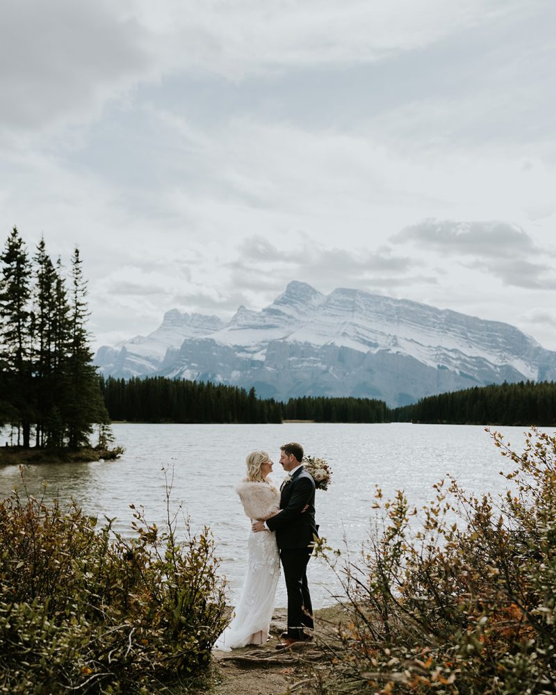 A bride and groom embrace by a scenic lake near Calgary, surrounded by trees and snow-capped mountains under a cloudy sky, creating a romantic and natural elopement atmosphere.