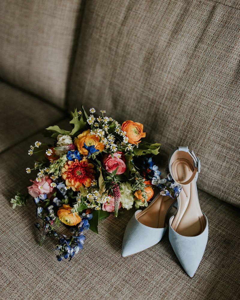 A colorful wedding bouquet of flowers and a pair of light blue pointed-toe shoes are placed on a beige fabric sofa, captured beautifully by a Canmore photographer.