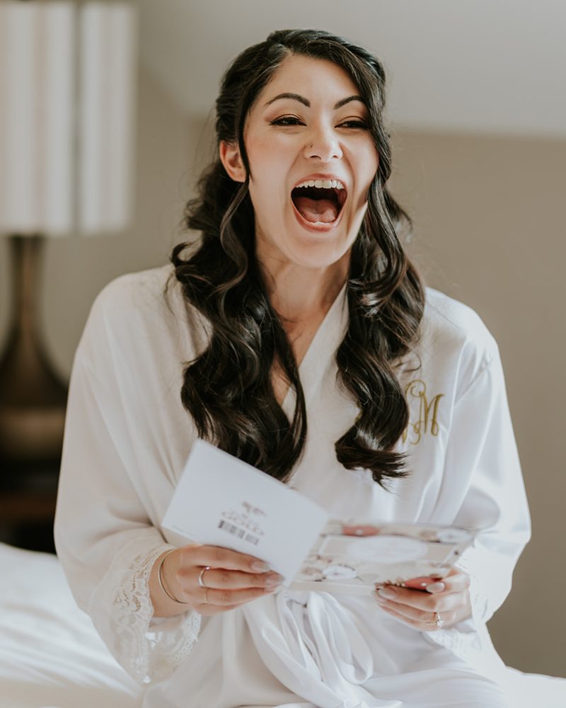 A woman in a white robe sits on a bed, laughing joyfully with her mouth wide open while holding an open card. Captured by a Calgary wedding photographer, the cozy scene features a lamp and a wooden bench in the background.