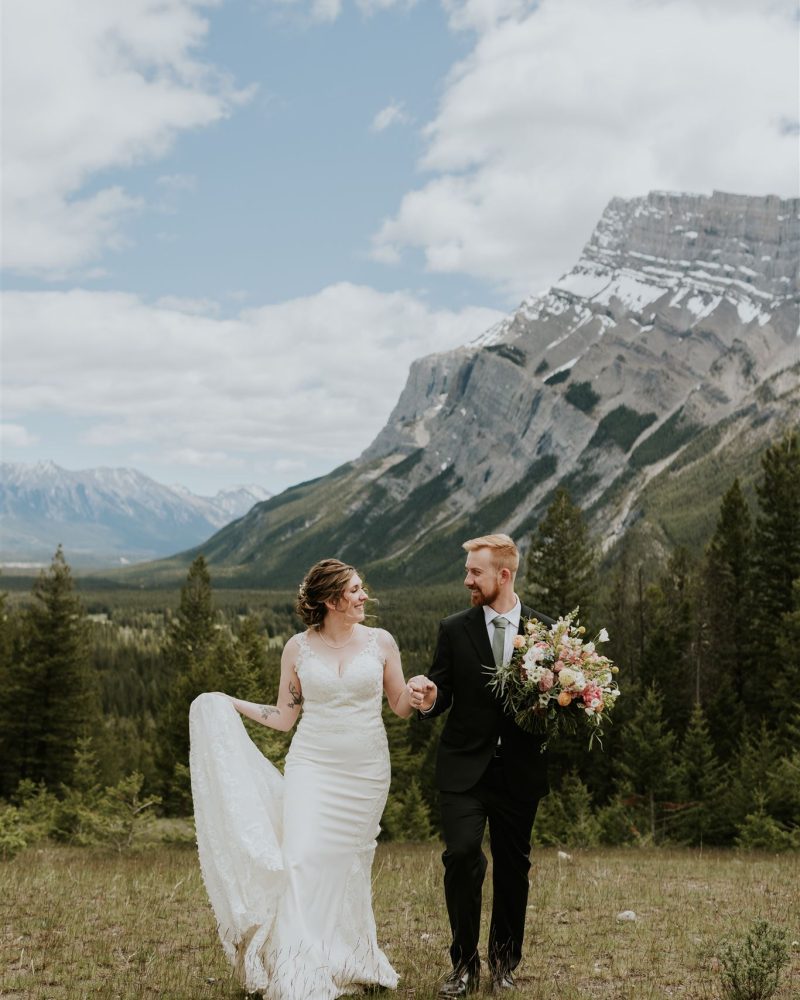 A bride in a white dress walks hand in hand with her groom in a black suit, holding a bouquet, as they celebrate their Canmore elopement against mountains, pine trees, and a partly cloudy sky.
