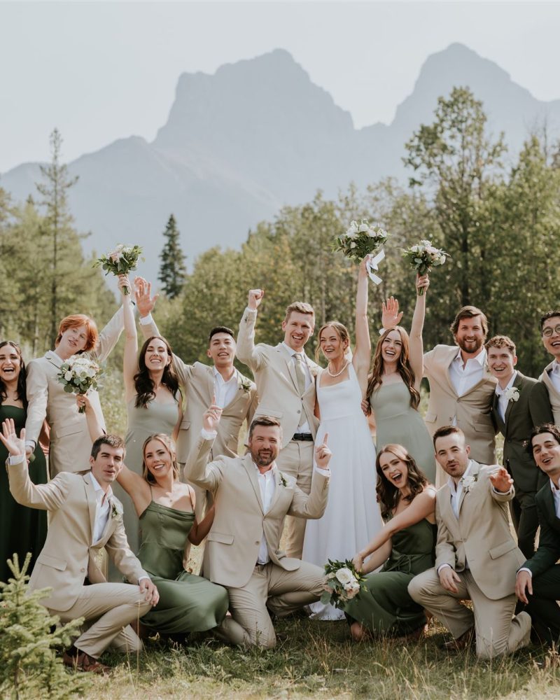 A joyful wedding party poses outdoors in Banff, surrounded by mountains and trees. The group, dressed in green and beige suits and dresses, raises bouquets and arms in celebration with the elopement photographer capturing the moment.
