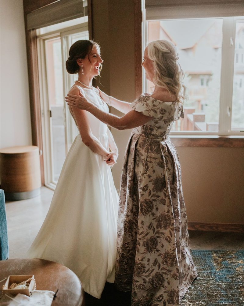 A bride in a white dress smiles as an older woman in a floral gown, possibly her mother, adjusts her necklace. Captured by a Calgary photographer, they stand in a softly lit room by large windows.
