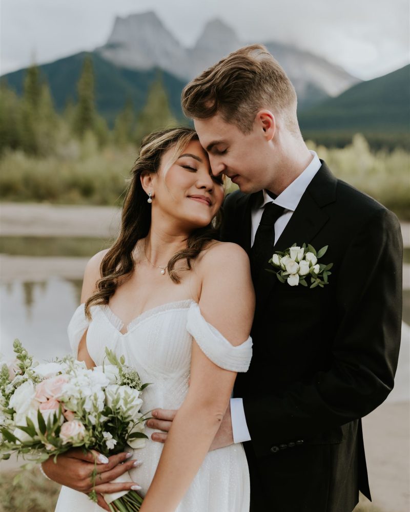 A bride and groom embrace outdoors, smiling softly. Captured by a Calgary wedding photographer, the bride holds a bouquet of white and pink flowers, while mountains and forest create a stunning natural backdrop.