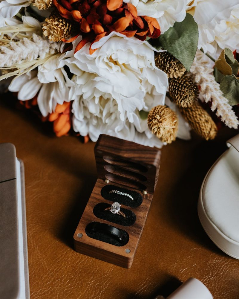 A wooden ring box holding a diamond engagement ring and band sits open on a brown surface beside a bouquet of white and orange flowers, a phone, round case, perfume bottle, and the essentials of any Calgary wedding photographer.