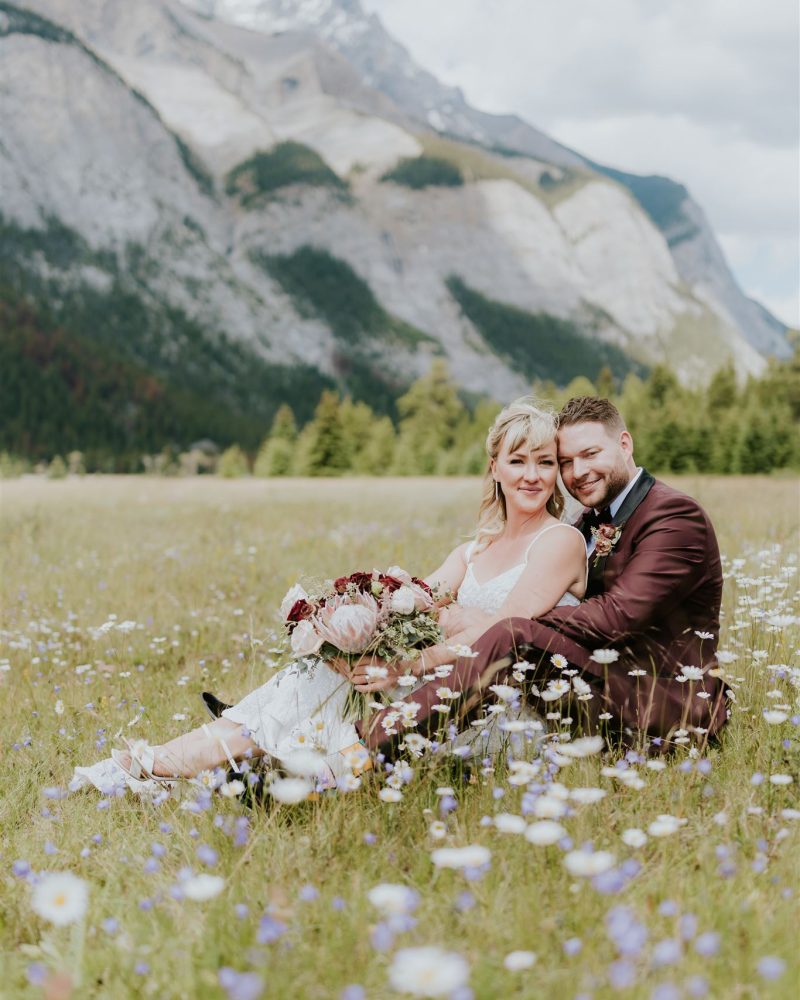 A bride and groom sit closely together in a wildflower field near Canmore, smiling, with mountains and trees in the background under a partly cloudy sky. The bride holds a bouquet of flowers on her lap, capturing their intimate elopement.