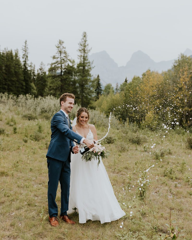 A bride and groom stand on a grassy field in Canmore, smiling and popping a bottle of champagne as it sprays. Mountains and trees form a scenic outdoor backdrop, perfect for an adventurous elopement.