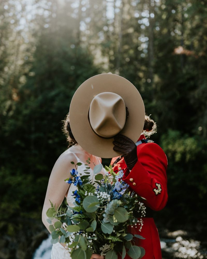 A couple poses outdoors in Banff among trees and sunlight, with one in a red uniform holding a wide-brimmed hat to hide their faces, and the other holding a bouquet of blue and white flowers—perfect for an intimate elopement.