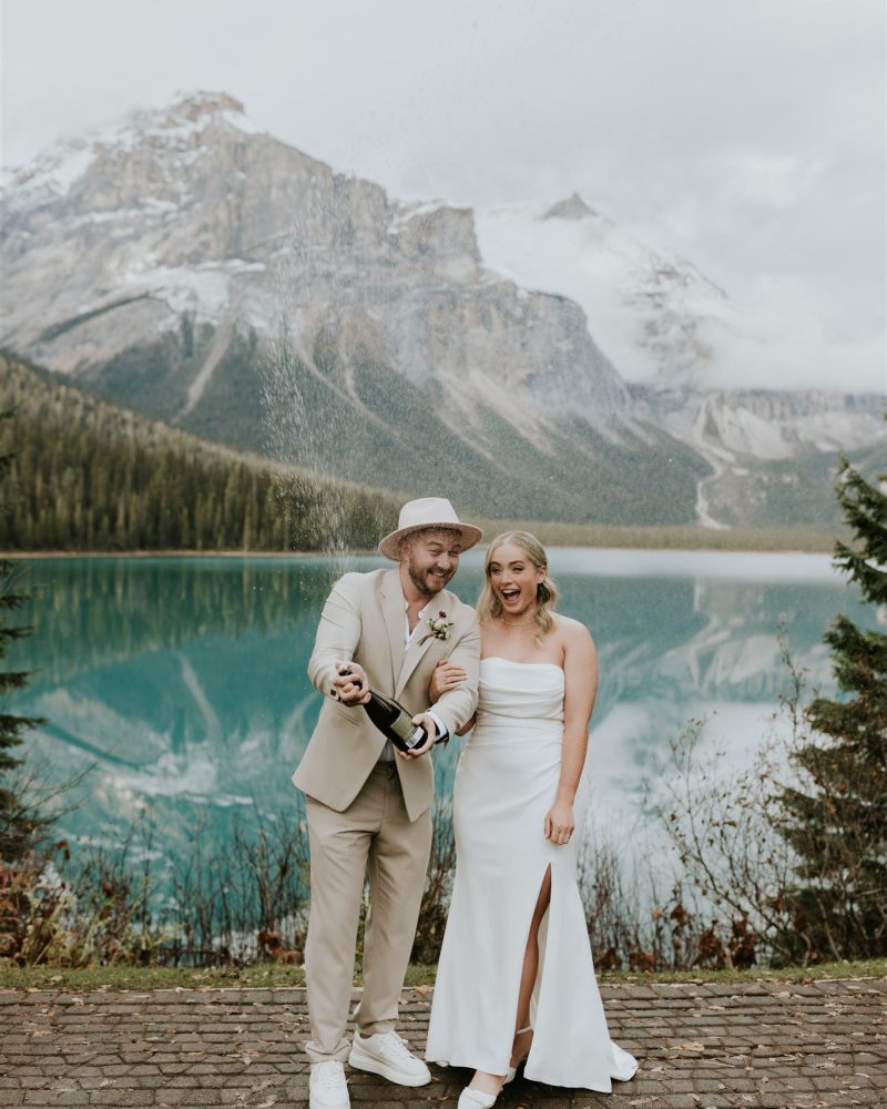 A couple in wedding attire stands by a turquoise lake and mountains near Canmore, smiling and popping a bottle of champagne outdoors on their Calgary wedding day.