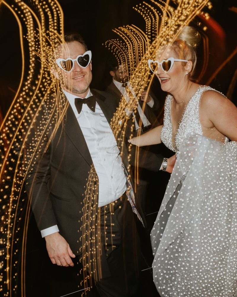 A couple dressed formally, both wearing heart-shaped sunglasses, smile and dance amid swirling, golden light trails at a festive event—captured perfectly by a Calgary photographer. The woman wears a sparkly dress and the man is in a tuxedo.