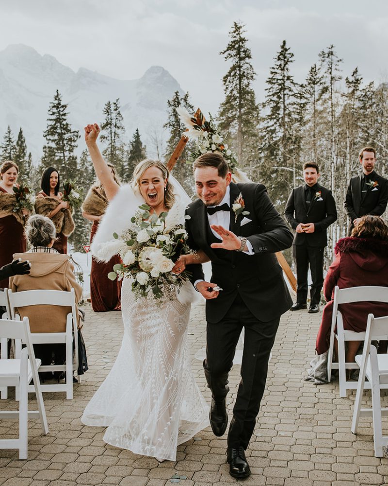 A joyful bride and groom walk down the aisle outdoors in Banff, celebrating with big smiles and raised arms, surrounded by guests seated in white chairs and a backdrop of snow-covered trees and mountains captured by their photographer.