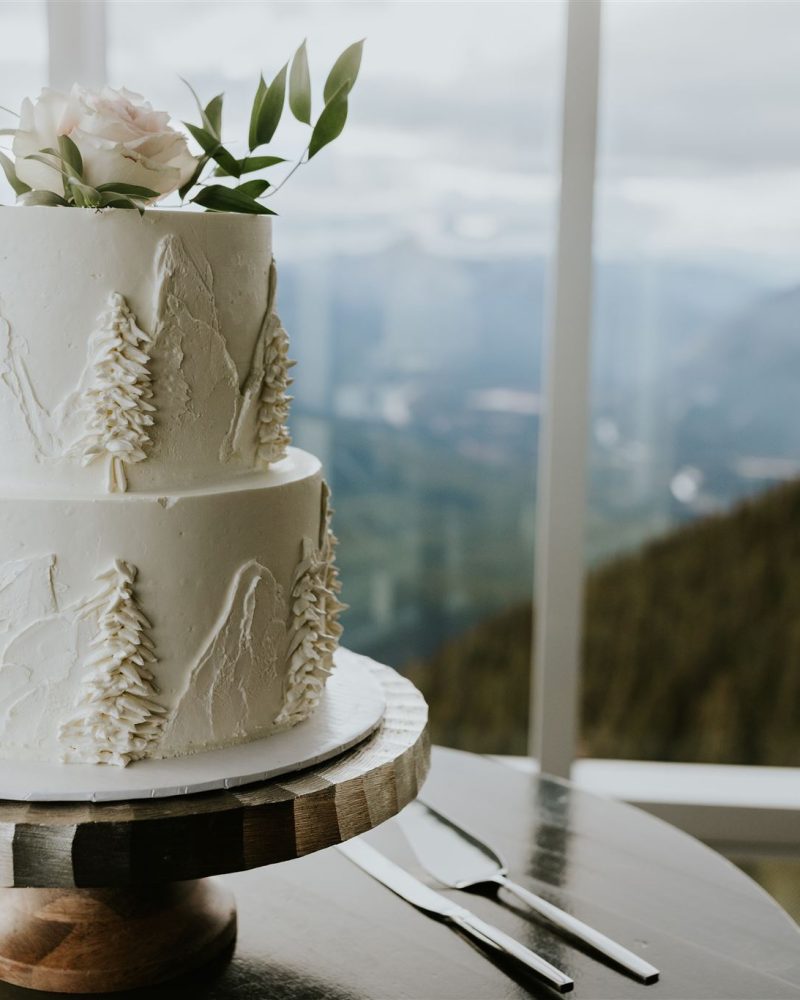 A two-tiered white cake decorated with piped trees and topped with a white flower and greenery sits on a wooden stand in front of large windows overlooking a stunning Banff mountain landscape—perfect for an intimate elopement.