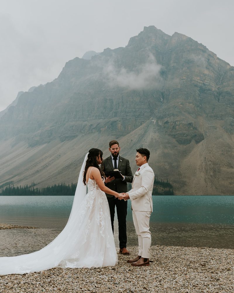 A couple holds hands during their wedding ceremony by a turquoise lake near Canmore, with a mountain and misty sky behind them. Their photographer captures the moment as an officiant stands between them on the rocky lakeshore.