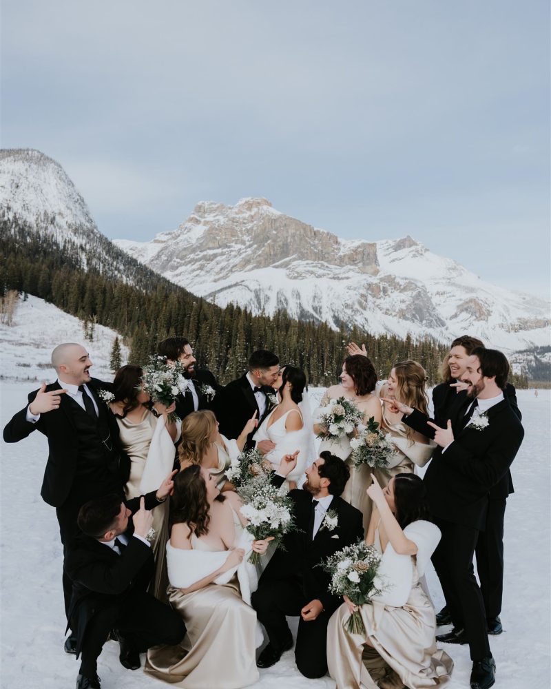 A wedding party poses playfully in the snow, with bridesmaids in champagne dresses and groomsmen in black suits, celebrating their Banff elopement surrounded by snowy mountains and pine trees under a clear sky.
