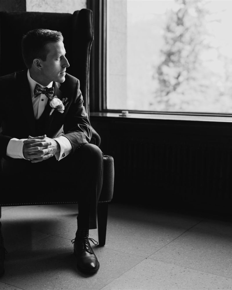 A man in a suit with a boutonniere sits on a chair, looking out a large window. Captured by a Calgary photographer, the black and white image highlights soft outdoor light and deep indoor shadows.